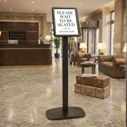 Black A4 floor-standing snap frame poster stand in a hotel lobby, displaying a please-wait-to-be-seated notice beside leather lounge seating.
