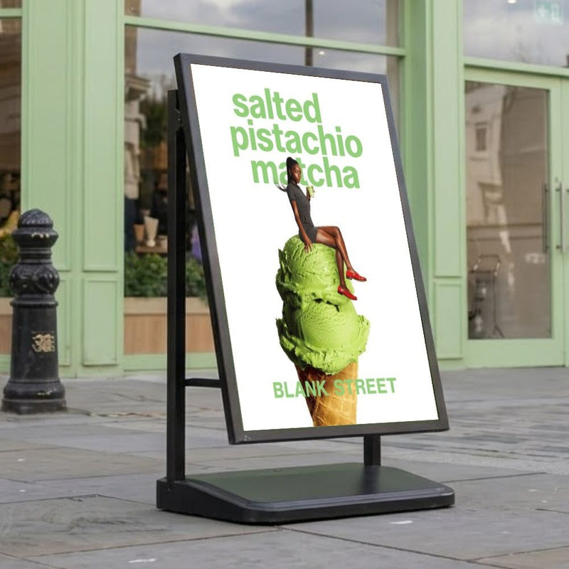 Black A1 single-sided waterbase pavement sign on a stone high street pavement outside a café with pale green shopfront, carrying an A1 promotional poster.
