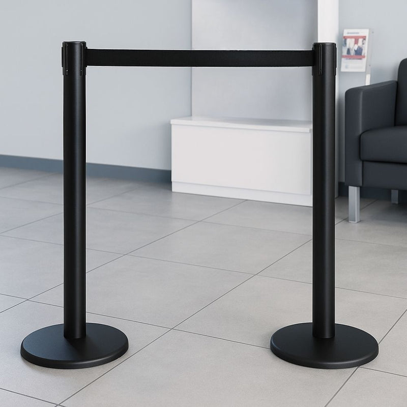 Pair of black barrier posts with black retractable webbing placed in a modern reception area with tiled flooring and minimalist seating.

