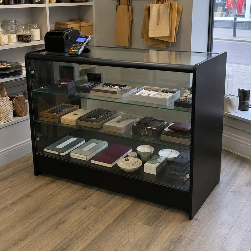 Black wooden glass retail display counter used in a boutique shop, featuring a glazed showcase front, internal display shelves and a flat top surface with card machine and till equipment for presenting jewellery, accessories, giftware and premium merchandise at the point of sale.
