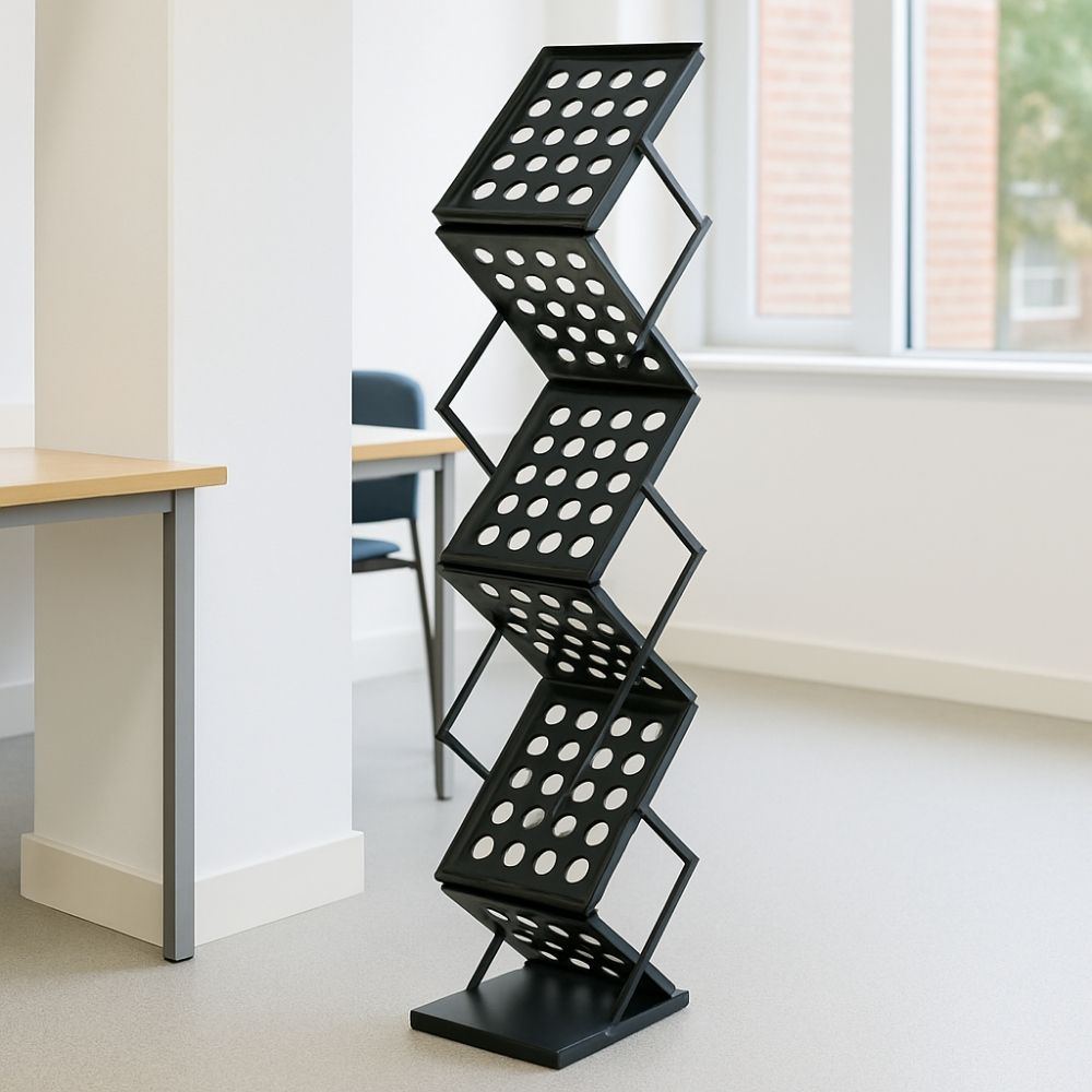 Black metal zigzag brochure stand with perforated shelves, positioned in a contemporary office space beside a wooden desk and window, ideal for leaflet and magazine display.