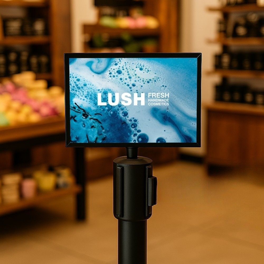A black queue barrier sign holder displaying a LUSH Fresh Handmade Cosmetics poster in a cosmetics shop.