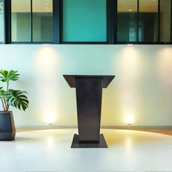 A modern black lectern positioned on a raised circular platform in a well-lit contemporary room. The lectern has a sleek, minimalist design with a wide top surface and a sturdy base. The background features a light-coloured wall with soft lighting and large windows framed with dark borders. To the left of the lectern, there is a potted green plant in a black planter. The setting appears to be a conference hall, lecture room, or event space.