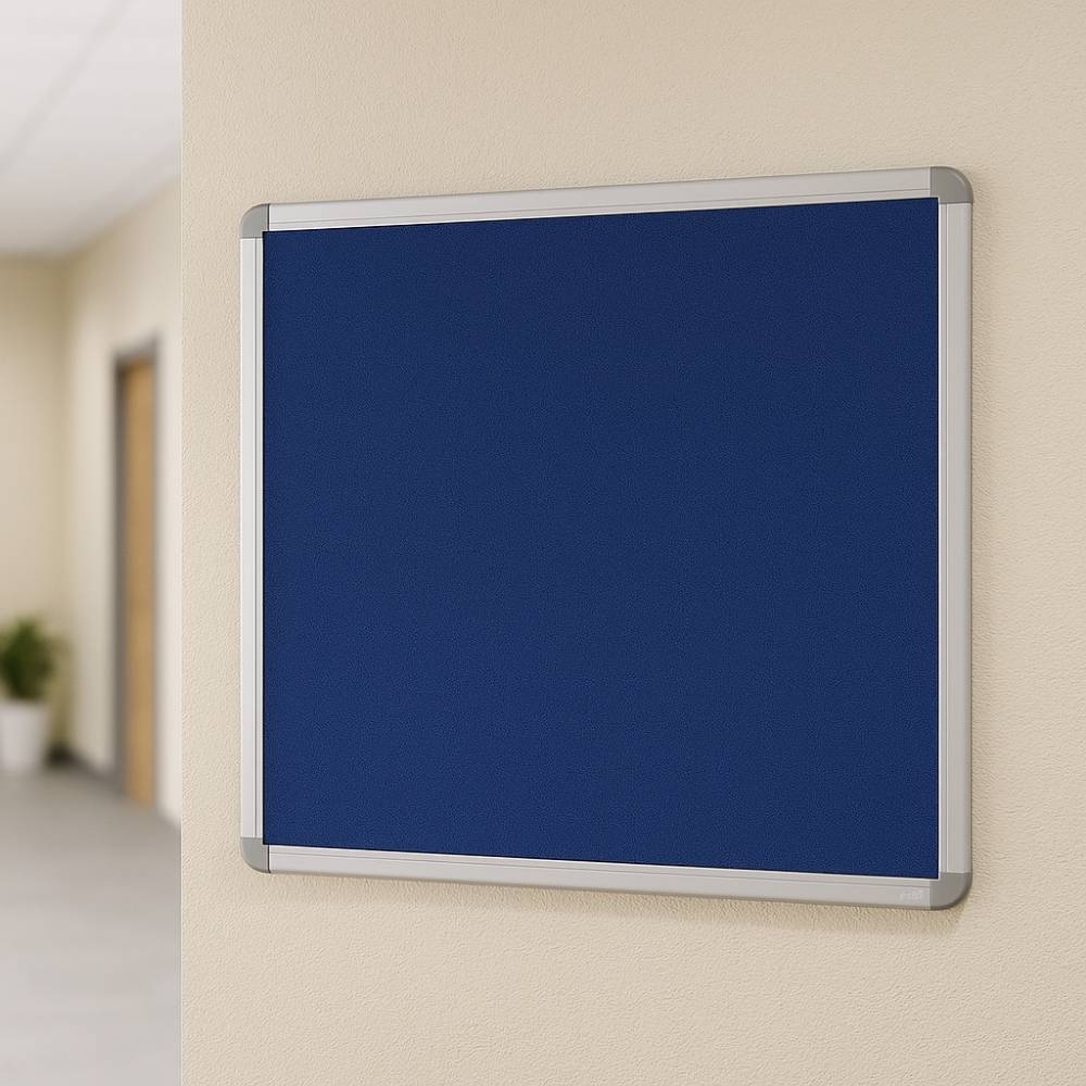 Blue felt noticeboard with silver aluminium frame mounted on a cream wall in a modern office or school corridor, with blurred hallway and plant in background.

