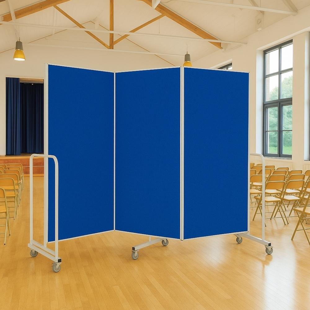 Mobile room divider with blue felt panels and silver frame on wheels, set up in a school hall with wooden floors and rows of yellow folding chairs.