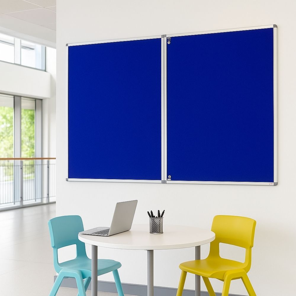Blue lockable noticeboard with aluminium frame mounted on a white wall in a bright classroom setting, shown above a round table with a laptop and two colourful chairs.
