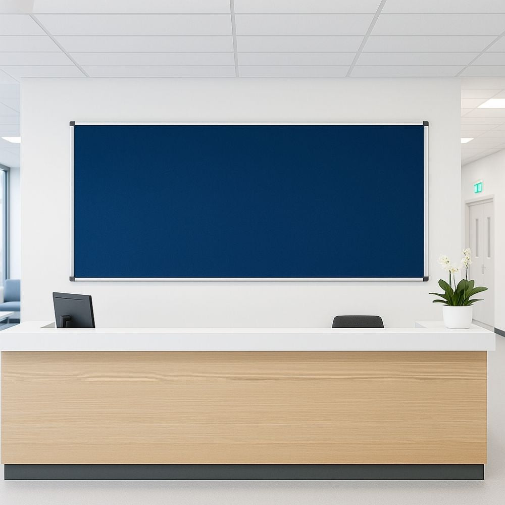 Deep blue noticeboard with silver frame mounted on white wall above modern wood and white reception desk in office setting
