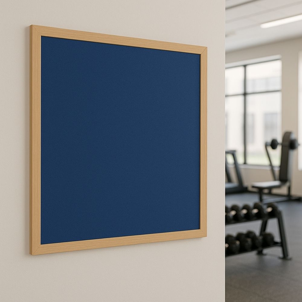 Blue noticeboard with a light wooden frame mounted on a wall inside a modern gym, with visible fitness equipment and natural light through large windows.