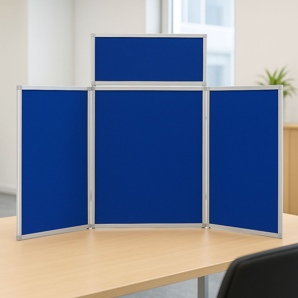 Blue tabletop display board with silver aluminium frame, set on a light wood desk in a bright office with a black chair and potted plant in the background.


