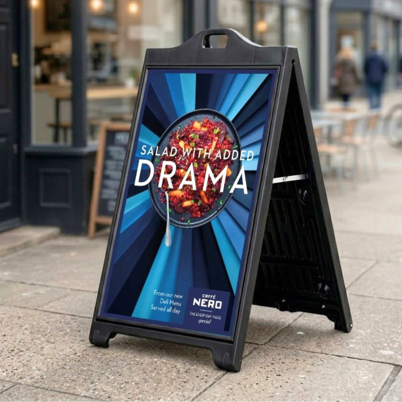 Outside a city café, the black A-board stands on the pavement with a full-colour poster on show, catching pedestrians walking past the shopfront.

