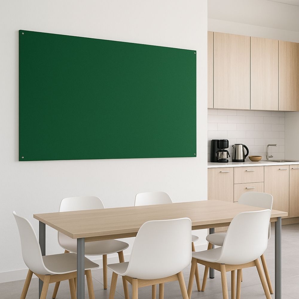 Green fabric noticeboard mounted on the wall of a contemporary office breakroom featuring a wooden dining table with white chairs and a kitchenette with light wood cabinets, a coffee machine, and a kettle.