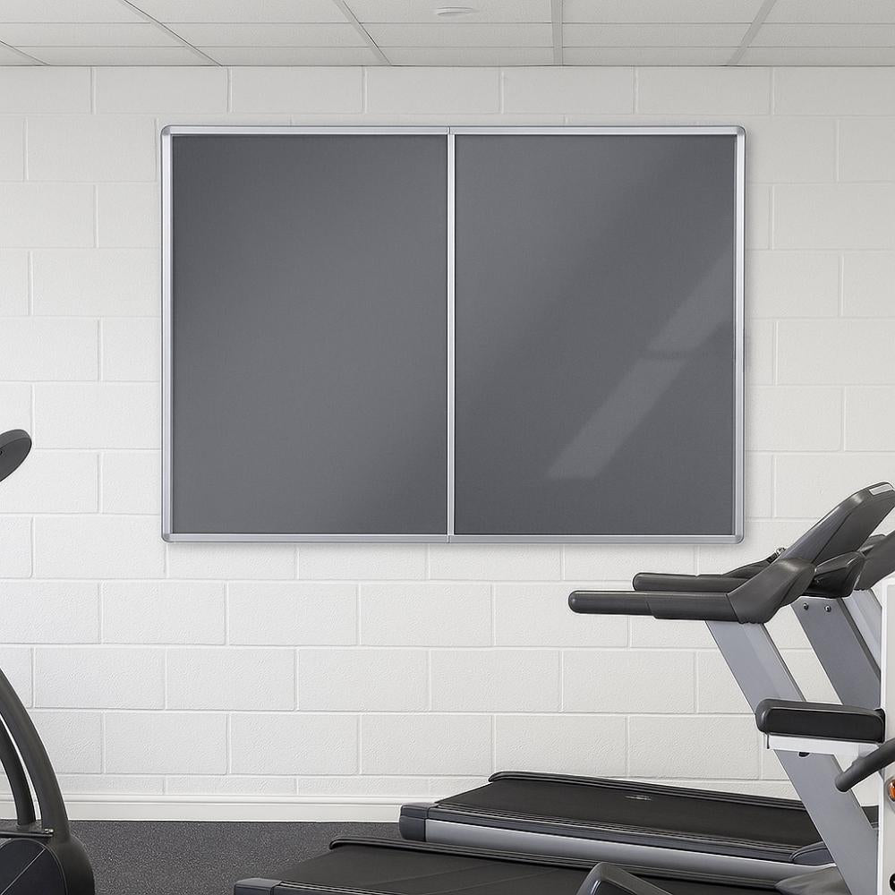 Grey double-door lockable noticeboard with silver aluminium frame mounted on a white brick wall in a gym, positioned above treadmills in a clean, well-lit exercise space.