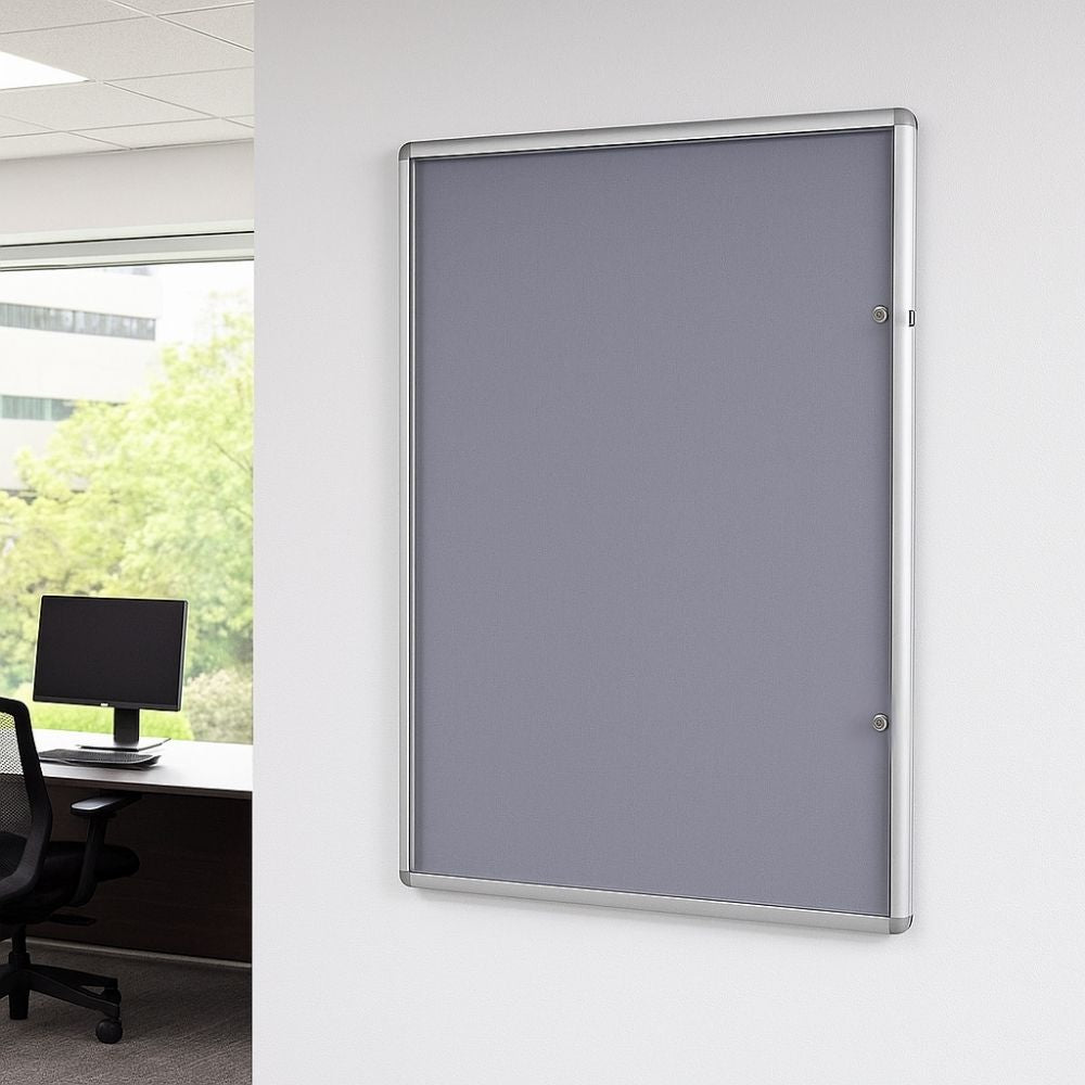 Grey lockable noticeboard with aluminium frame securely mounted on a white office wall beside a window and black mesh chair.

