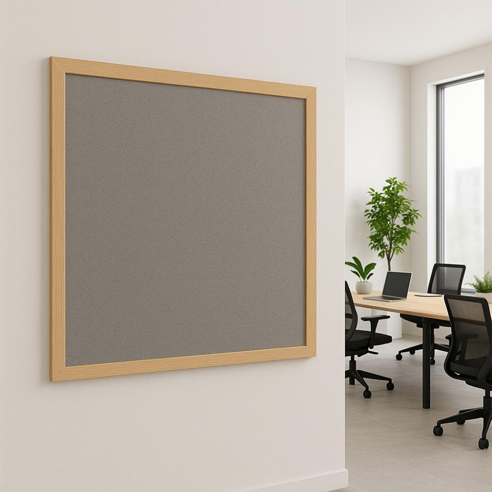 Wall-mounted grey noticeboard with a light wooden frame in a modern office meeting room, featuring black chairs, laptops, and indoor plants near large windows.