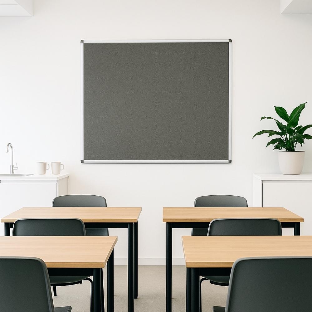 Grey classroom pinboard with aluminium frame on white wall, positioned above wooden desks and black chairs, near plant and sink unit