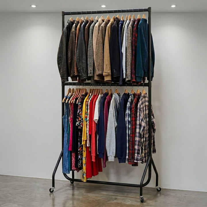 Black two-tier clothes rail on wheels holding a large assortment of jackets, shirts and dresses in a minimalist indoor space.