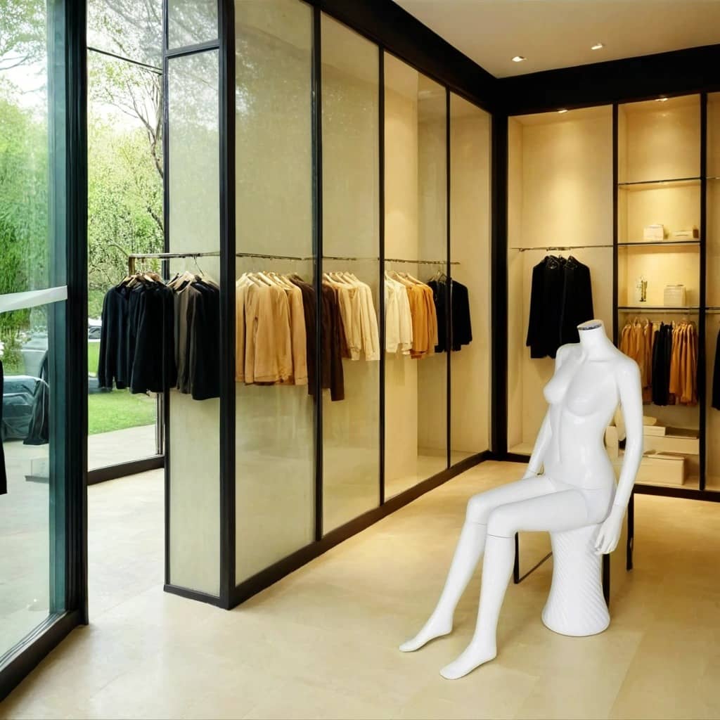 A modern and sophisticated clothing boutique with floor-to-ceiling glass panel walls, offering a view of lush greenery outside. Inside, neatly arranged garments in neutral and earthy tones hang on minimalist black clothing racks. A headless, glossy white female mannequin sits gracefully on a sculptural white stool, positioned to complement the store's sleek and contemporary design.