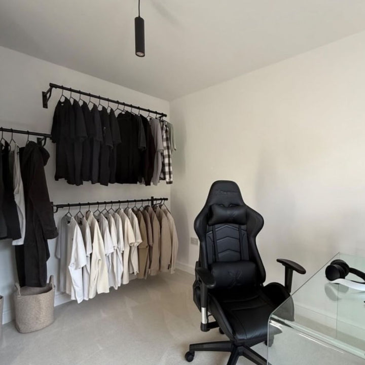 A modern home office featuring black wall-mounted clothes rails displaying monochrome clothing, a black ergonomic gaming chair, and a glass desk with headphones.