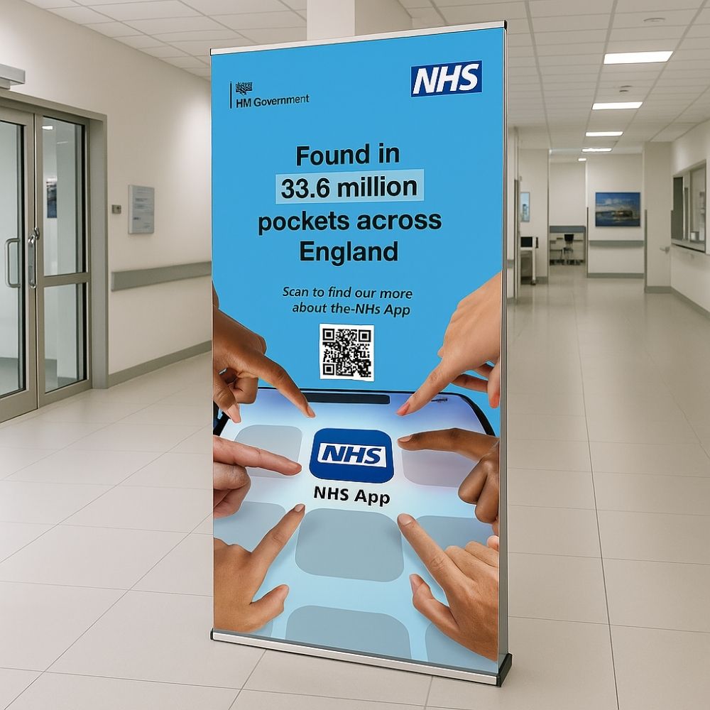 NHS App promotional roller banner displayed in a hospital corridor, highlighting app availability and download access for 33.6 million users across England.