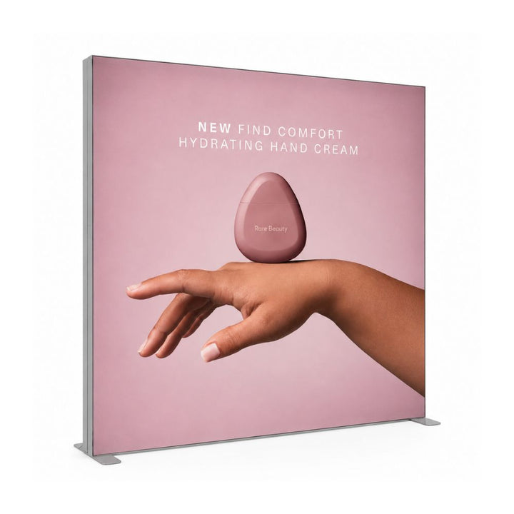 Backlit advertising display promoting Rare Beauty hydrating hand cream with minimalist pink cosmetic branding and hand model imagery.