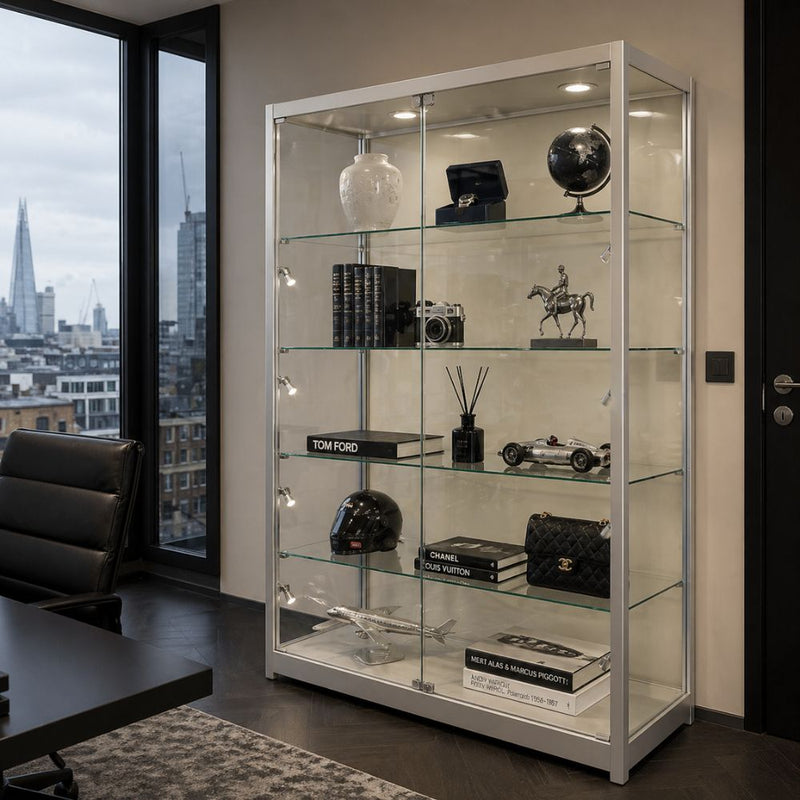Silver aluminium lockable glass display cabinet with LED lighting showcasing decorative items and accessories in a modern office interior with city view.