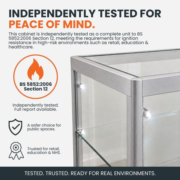 Silver aluminium glass counter display cabinet highlighting independent BS 5852:2006 Section 12 ignition resistance testing for retail, education and healthcare environments.