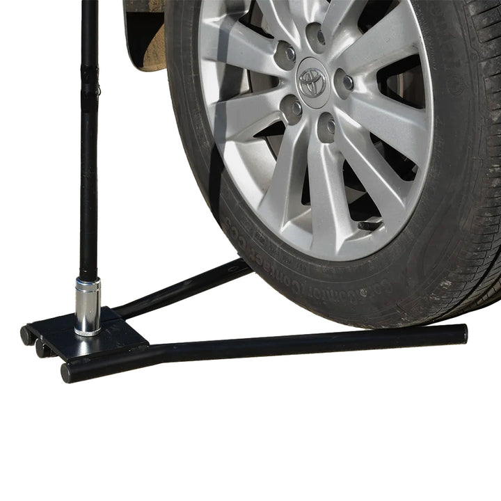 Close-up of the steel drive-on car base with a vehicle tyre anchoring a teardrop flag on a hard forecourt surface without fixings or ground damage.
