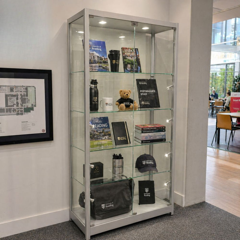 Prospectuses, a water bottle, mug, cap and messenger bag fill five lit levels of a silver framed cabinet in a university foyer, doors locked at the centre.
