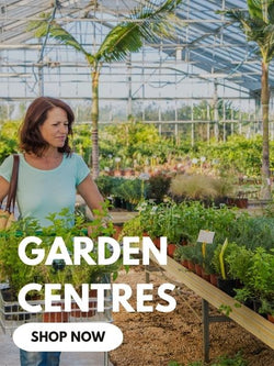 Woman pushing a trolley filled with potted plants inside a bright garden centre greenhouse filled with greenery.

