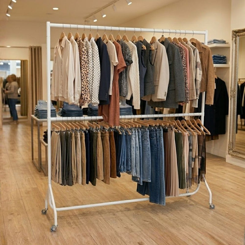 Blouses and jackets fill the upper bar while trousers and jeans hang below, the white two-tier rail set on the shop floor of a womenswear boutique.
