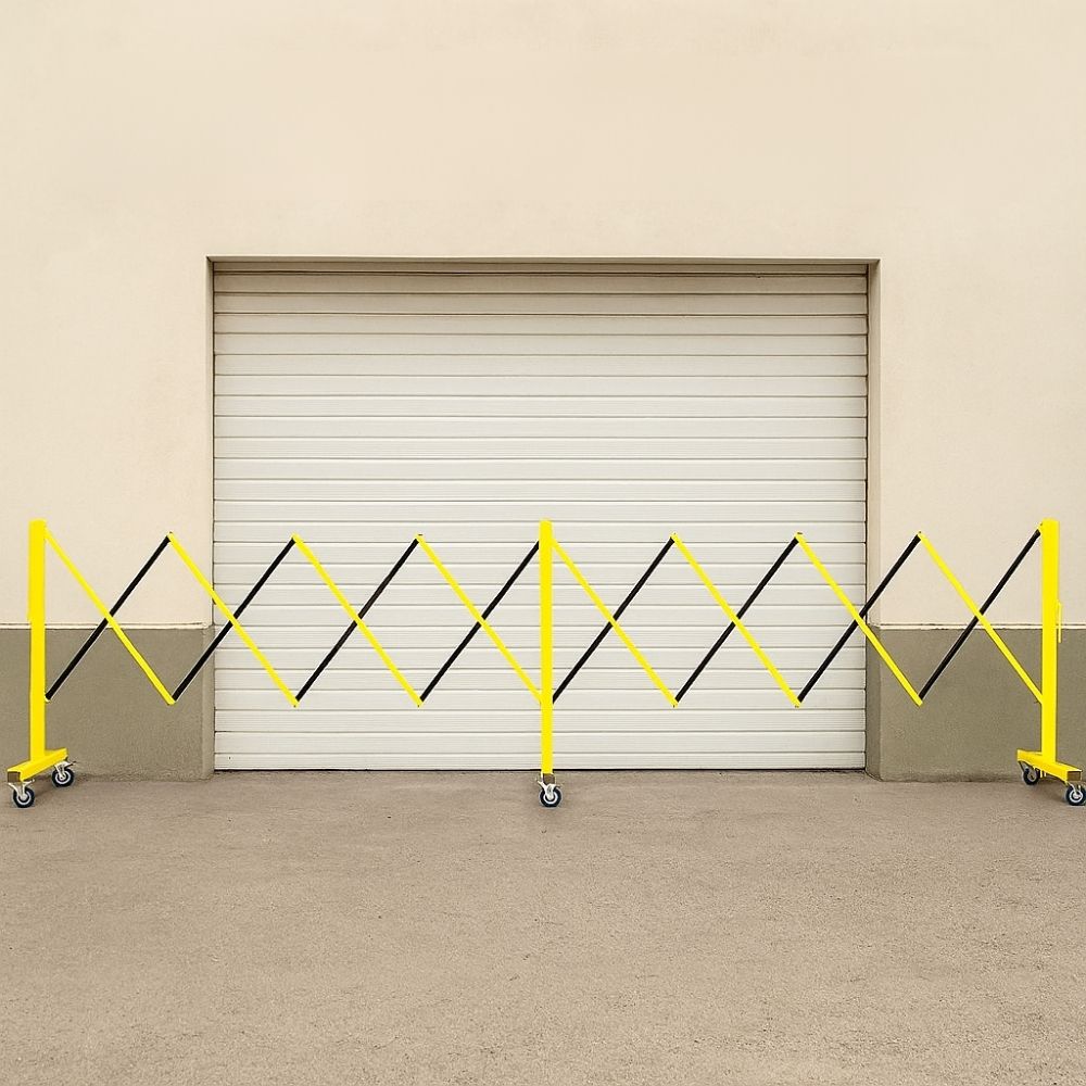 Yellow and black expandable safety barrier on wheels placed in front of a closed roller door outside a building.