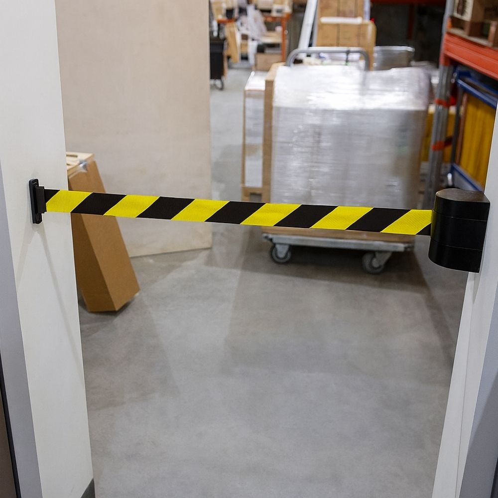 Wall-mounted retractable safety barrier with yellow and black striped webbing stretched across a warehouse doorway with pallets and trolleys visible.